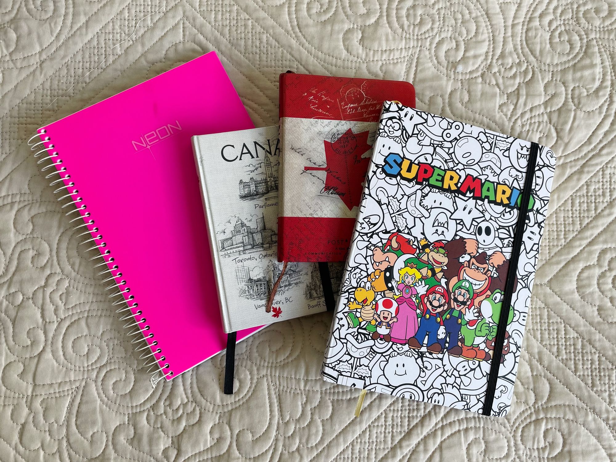 Um caderno rosa, seguido por dois pequenos cadernos com temática canadense e um caderno branco com capa do Super Mario World.